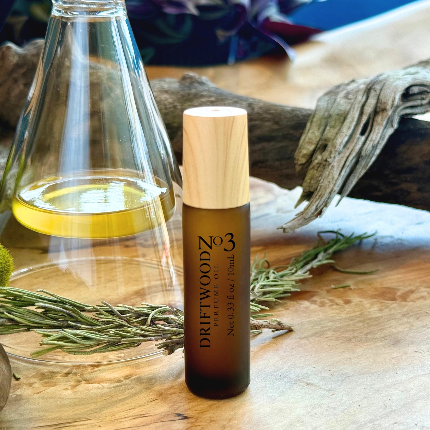 Driftwood No. 3 frosted amber perfume oil bottle on a wooden surface with rosemary, driftwood and a glass perfume sniffer container.